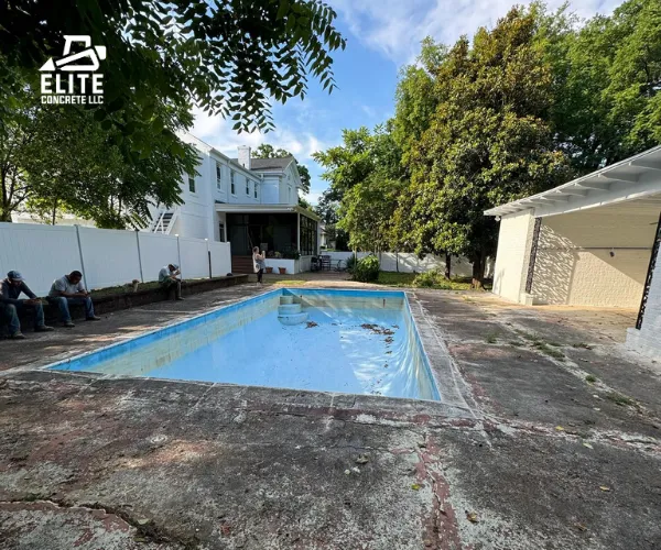 A ground-level view of an older inground pool surrounded by a cracked and weathered concrete deck