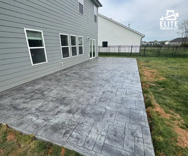 A newly installed decorative stamped concrete patio with a dark grey ashlar slate pattern bordering a residential backyard lawn.