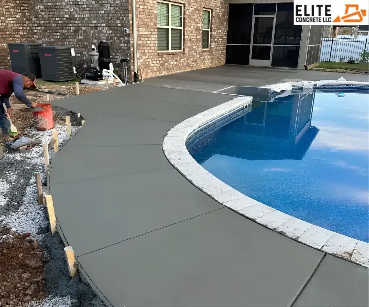 Wide shot of a newly poured grey concrete deck surrounding a curved inground swimming pool next to a brick house.